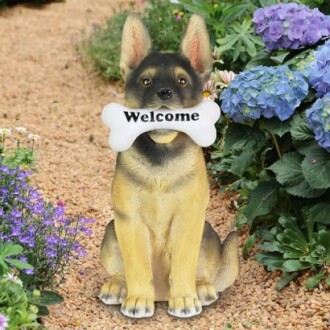 Dog statue holding 'Welcome' bone in garden with flowers.